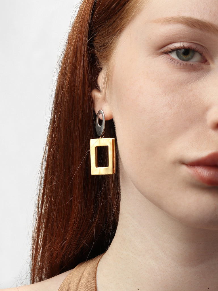 woman wearing dangle stud earrings with silver oval motif on the top and gold square motif on the bottom