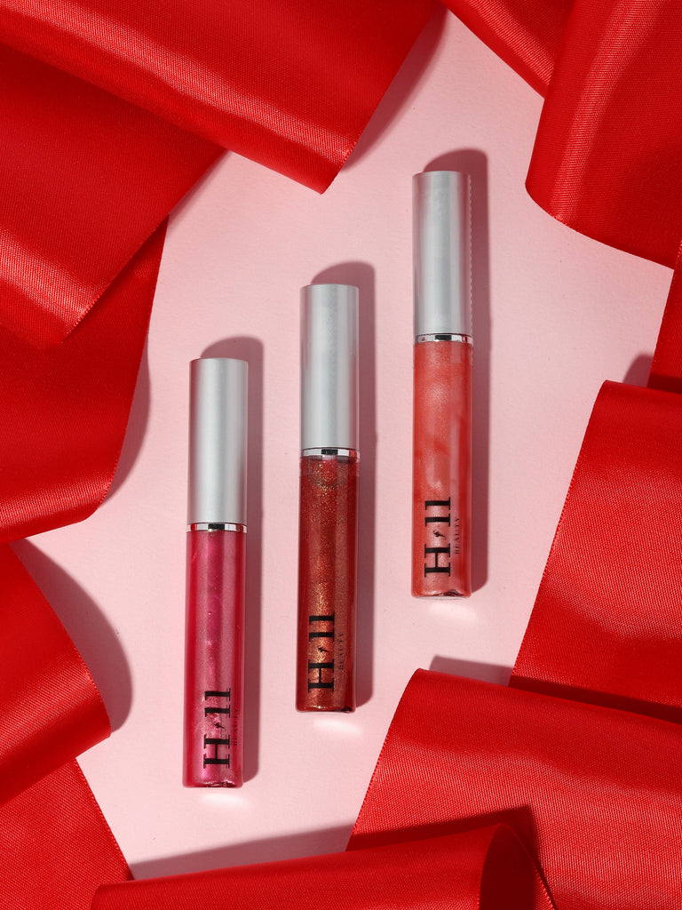 three lip glosses resting against a pink background surrounded by red ribbon