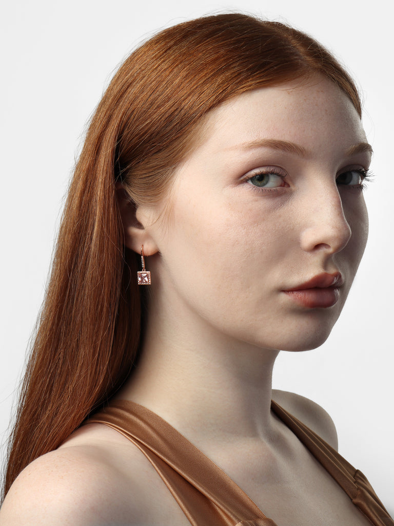 woman wearing rose gold dangle hook earrings with large square, pink gem surrounded by small clear-colored crystal gems