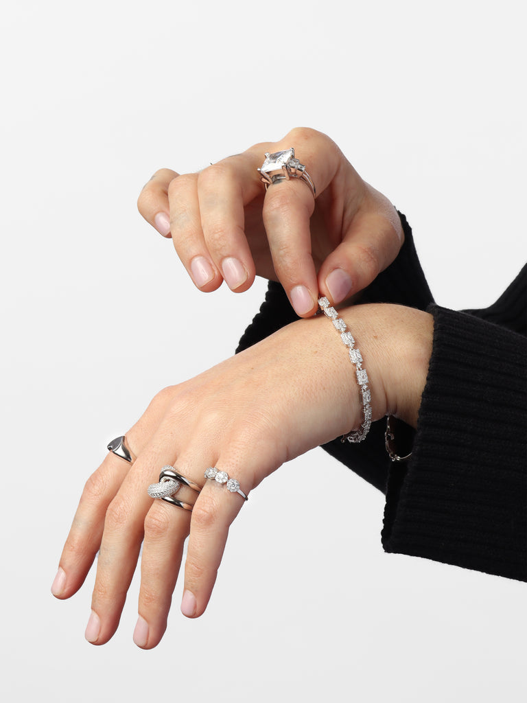 woman's hands wearing silver bracelet with rectangular link motif. In the center is a rectangle shaped, clear-colored gem outlined by small round clear-colored crystals.
