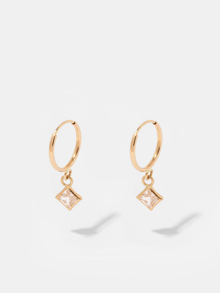 "Image description: A pair of earrings featuring square drop pendant crystals housed in mini solid 14K yellow gold hoops designed to dangle and sparkle with subtle movements. Each earring consists of 14K solid yellow gold with a square-cut cubic zirconia stone. The hoop measures 11.6mm in diameter, and the stone measures 5.7mm in width."